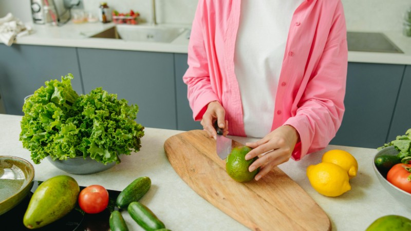 Person in pink shirt chopping vegetables on a wooden board, surrounded by fresh produce., tags: für - pexel