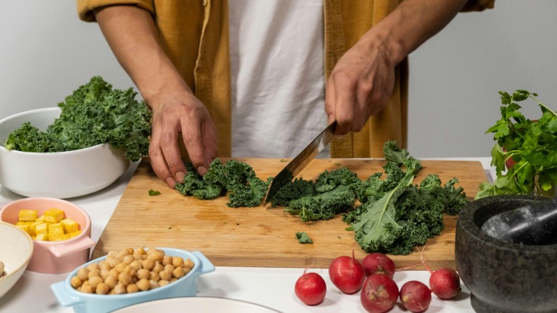 A person chops fresh kale surrounded by vibrant vegetables for a healthy meal preparation in a kitchen., tags: abendessen der und - pexel